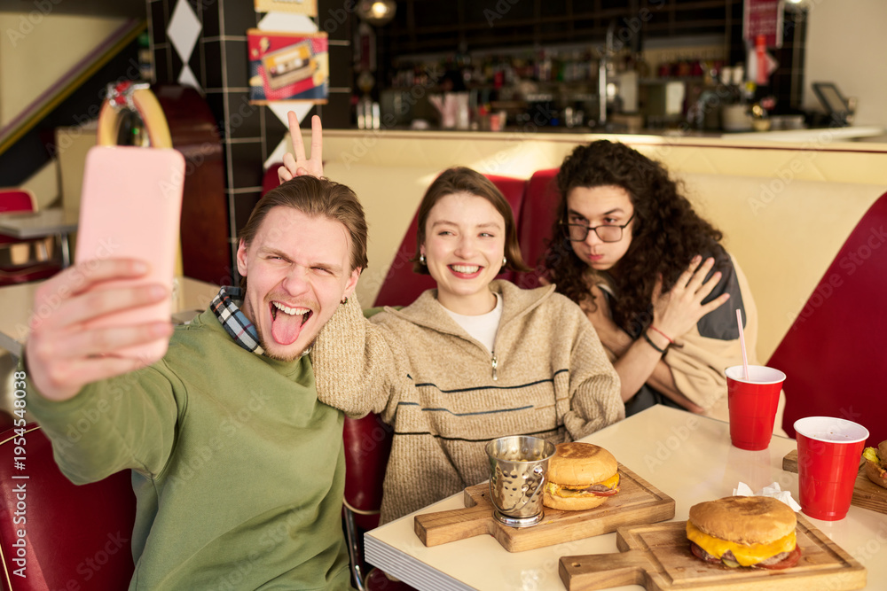 Obraz premium Three gen Z friends smiling and posing, taking selfie together in diner with burgers