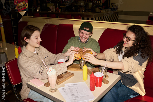 Three young friends sitting in retro diner booth sharing burgers and milkshakes, smiling and enjoying meal together, food and drinks on table