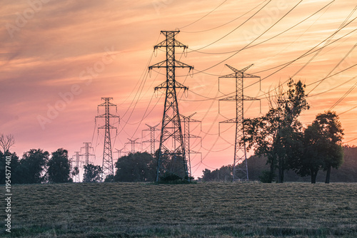 Linie wysokiego napięcia i maszty energetyczne na tle malowniczego zachodu słońca. Infrastruktura energetyczna na polu w ciepłym, pomarańczowym świetle wieczornym.