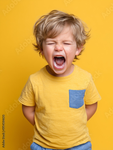 Cute little child with blond hair screaming loudly against a bright yellow background, expressing strong emotion and childhood tantrum.