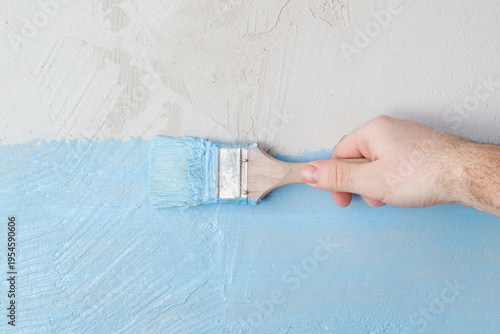 Young adult man hand holding brush and applying blue waterproofing coating on concrete wall or floor before lying tiles at bathroom. Closeup. Repair work of home. Front view.