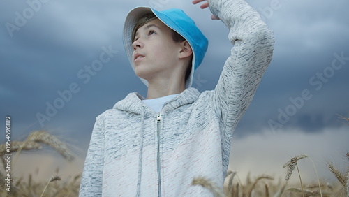 Bild auf Leinwand Teenage Boy in Blue Cap Standing in Wheat Field with Cloudy Sky, have fun and en