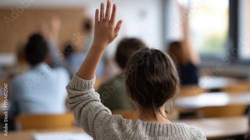 Female student raising hand in classroom filled with other students, wooden desks and papers visible, bright natural light coming through windows