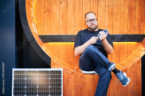 Bearded man connecting smartphone to photovoltaic solar panel. Integration of sustainable renewable energy into everyday life, demonstrating practical use of solar power for charging devices.