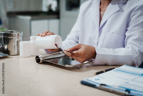Pharmacist or lab technician in white coat counts red tablets from pill bottle onto a tray at a laboratory desk with documents nearby.