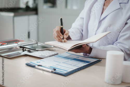 Doctor in white coat writing notes on clipboard at clinic desk with medical forms, pen, keyboard and medicine bottle; healthcare documentation and prescription concept.