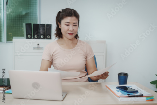 Professional woman reviewing paperwork at office desk with laptop, coffee, smartphone and folders, modern workplace, focused expression, business administration concept.