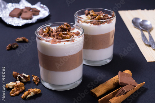 Delicious dessert served in two glasses with layers of cream jelly, chocolate jelly, nuts, and spices on a dark table