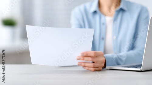 Person holding a blank certificate paper in hand while sitting at a desk with a laptop, showcasing a modern workspace with soft lighting and minimalistic decor in the background