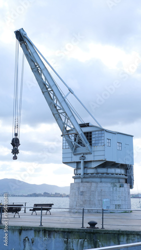 A vintage industrial harbor crane sits dormant on a stone plinth, its metal boom angled toward a cloudy sky 