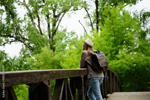 Female tourist rests amidst picturesque nature, leaning against large wooden railing. Traveler rests on rustic bridge crossing lush green forest. Concept of eco-trail, uniting people and nature.