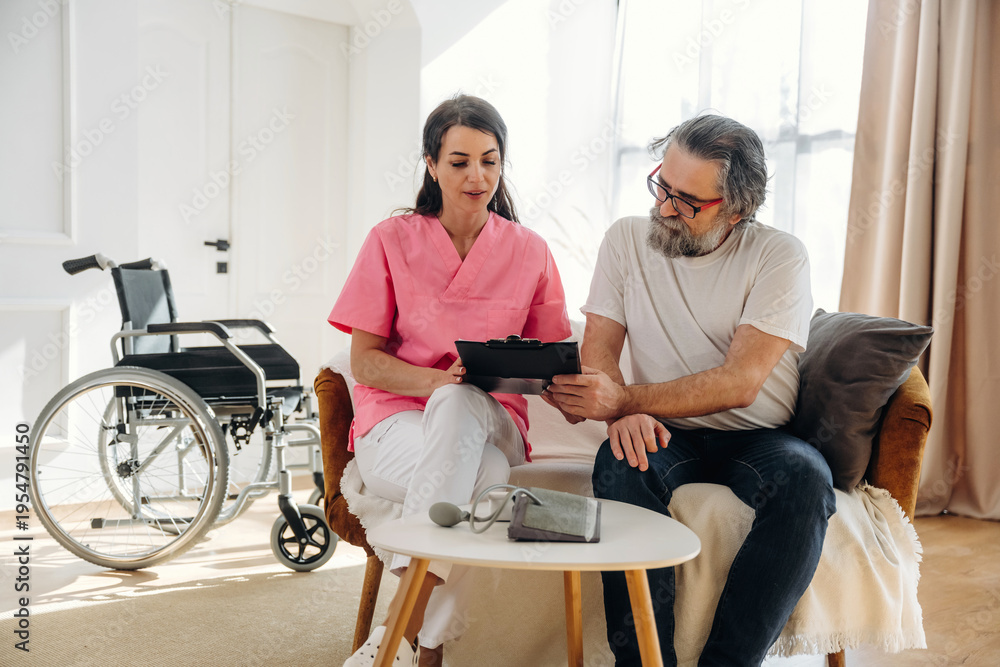 Fototapeta premium Female healthcare professional in pink scrubs discusses treatment options with senior man in casual attire, wheelchair positioned nearby in a well-lit living room setting