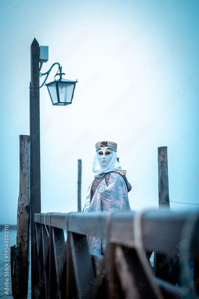 Fototapeta premium Venice, Italy - February 13, 2026: Person wearing ornate mask and costume stands on wooden pier with lantern, foggy atmosphere, Venice Carnival celebration in the background