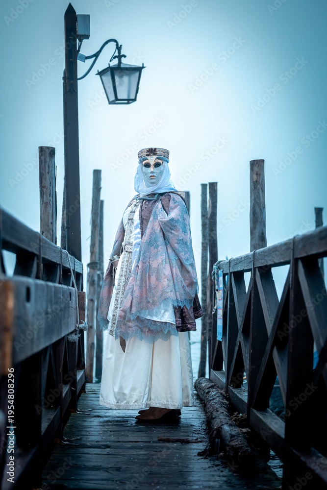 Fototapeta premium Venice, Italy - February 13, 2026: Woman in elaborate costume and mask stands on a wooden pier during Venice Carnival, lantern and fog creating a mystical atmosphere