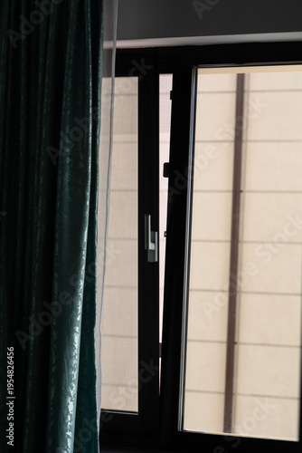 A PVC window in ventilation mode, with green curtains on the side. Selective focus, shallow depth of field. View from the window onto the wall of a neighboring building.