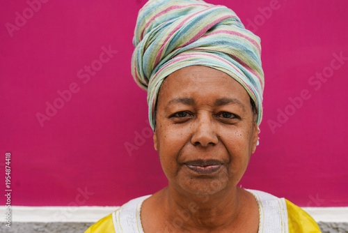 African senior woman looking at camera outdoor with pink wall in background - Portrait of elderly person wearing traditional clothes - Copy space