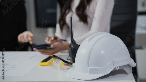 White safety hard hat with walkie-talkie and safety glasses on desk in office, blurred engineers discussing plans in background; workplace safety and construction project management concept.