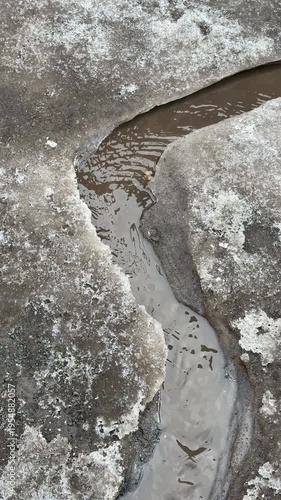 Spring ice melting, water flowing through cracks in concrete, urban thaw, nature breaking through manmade surfaces, dynamic small stream, urban micro-river, seasonal change, close-up nature flow