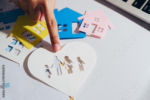 Paper cutouts of houses in various colors surround heart shaped cutout depicting family holding hands. finger points to family cutout, symbolizing love and connection