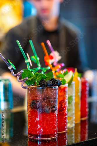 Colorful cocktails lined up on a bar counter during a busy evening. Five drinks with bright colors sit on a bar counter while a bartender prepares another order in the background.