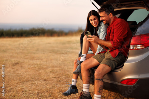 Couple sitting in a car trunk using smart phone and relaxing in nature