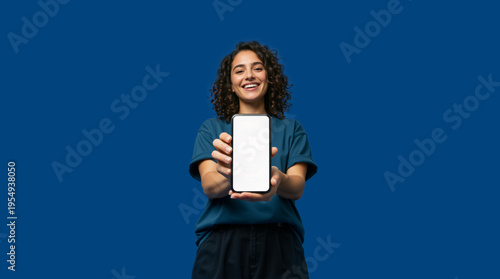 Smiling young woman showing blank smartphone screen on blue background. Happy female holding mobile phone for app mockup or digital advertisement. Copy space for text