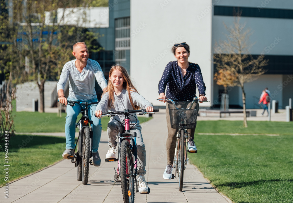 Obraz premium Happy family with a young daughter riding bicycles on a paved path in a sunny park as a symbol of active lifestyle and healthy outdoor recreation for parents and children during a family weekend trip