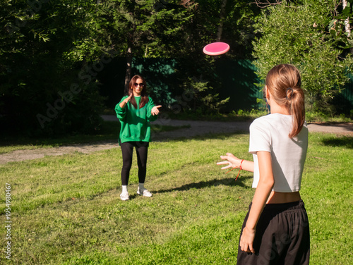 Mom and daughter playing frisbee in a sunny park. Enjoying outdoor physical activity with flying disc. Active lifestyle, leisure, friendship and sports game concept in summer nature.