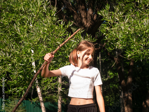 Young happy girl playing with stick in summer park. Child having fun in forest during sunny day. Playful kid enjoying nature activity outdoors. Cheerful childhood lifestyle concept.