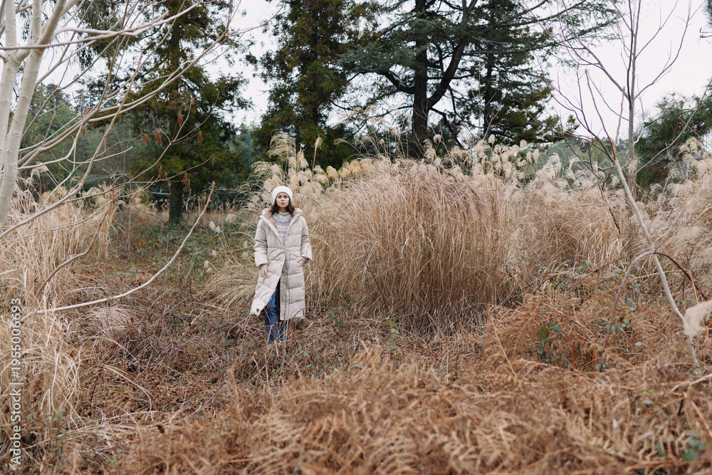 Fototapeta premium Winter scene with a lone person in a light coat standing among tall dried grasses in an open field, calm and natural, highlighting solitude and outdoor exploration.