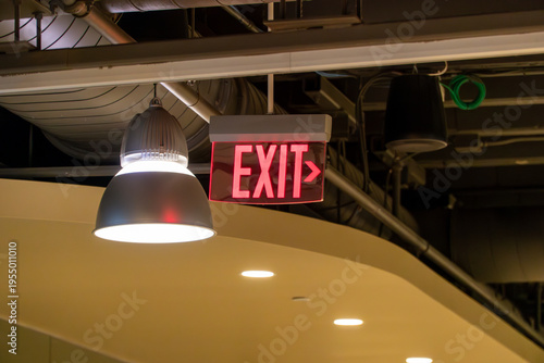 Photos Bright red illuminated EXIT sign with arrow, hanging prominently from a commercial building ceiling next to an industrial pendant light fixture, indicating emergency egress route