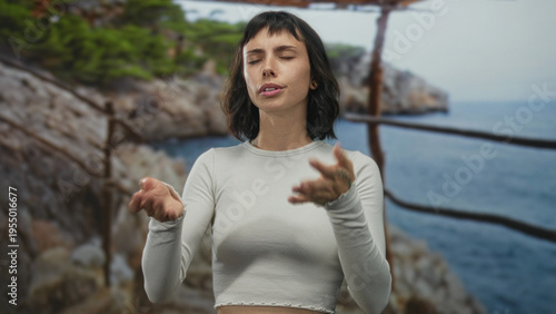 Slika na platnu Young hispanic woman fans face with hands beside a rugged rocky coast railing in forest; relief from heat