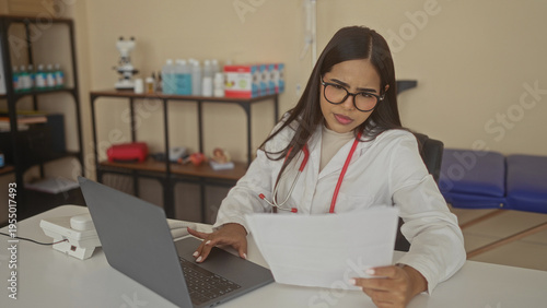 Foto Doctor woman wearing stethoscope and glasses holds paper and types on laptop at desk in clinic building; focused concentration