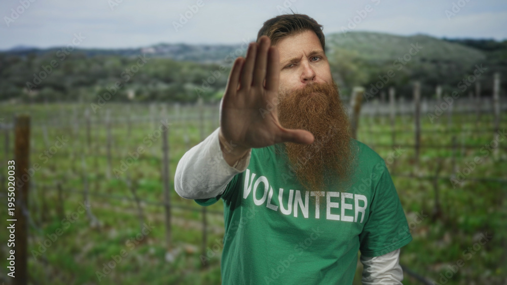 Fototapeta premium Man wearing green volunteer shirt raises hand in stop gesture in a countryside field; safety awareness.