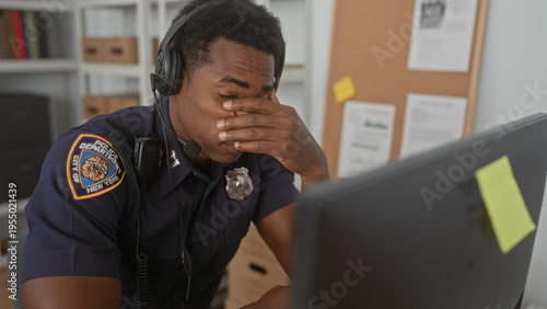 Foto Man police officer in uniform wearing headset covers face with hand at desk by computer monitor and corkboard in building; stress