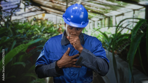 Fotografie Man in blue hardhat coughing and clutching chest among plants inside a plant filled building builder visible; safety concern