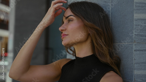 Hispanic woman hand on chin as hair blows gently while leaning against tiled wall in busy street; reflection.