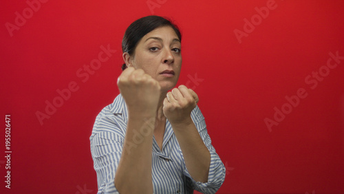 Φωτογραφία Middle aged woman in striped shirt with bare fists raised in fighting stance in red studio setting; defiance