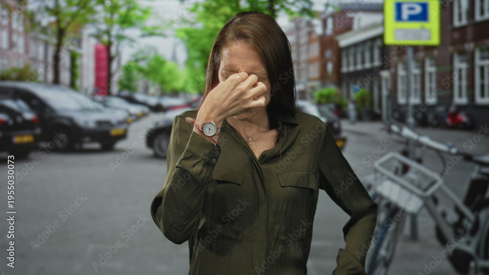 Fototapeta premium Young hispanic woman pinches nose with watch visible on a city street near parked cars and a bicycle rack and trees by buildings; disgust.
