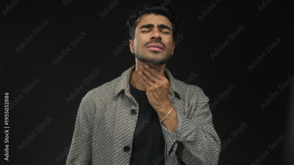 Fototapeta premium Young hispanic man hand on neck with pained expression in studio with black backdrop; discomfort.