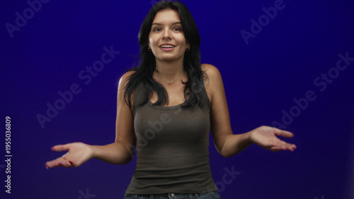 Hispanic woman wearing a brown tank top holds open palms and smiles broadly in blue studio setting; happiness.