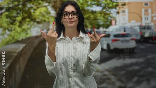 Woman in white striped shirt and glasses flipping both middle fingers on sunny street by parked cars and green trees; defiance.