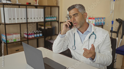 Bilde på lerret Man doctor in white coat holds phone to ear and gestures with hand over laptop at desk in medical office building; concerned care