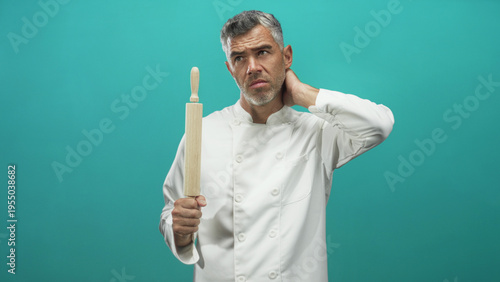Принти на полотні Chef man in white coat with grey hair holding wooden rolling pin, rubbing his neck and squinting in studio; quiet weariness