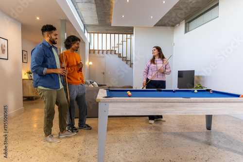 Diverse friends playing pool around blue felt table in modern home rec room, holding drinks