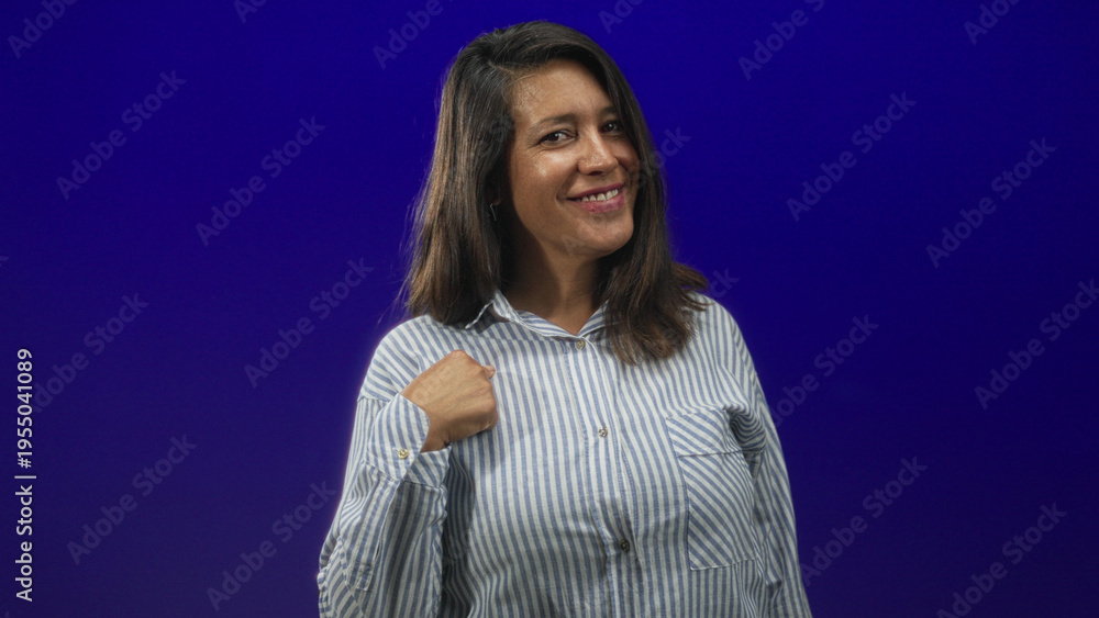Obraz premium Woman points fist to chest in studio, striped button shirt visible with confident smirk toward camera; confidence.