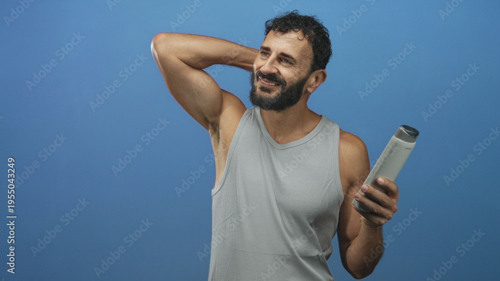 Fototapeta premium Man holding shampoo bottle, hand behind neck showing bare arm in blue studio; self care grooming confidence.