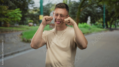 Young man rubbing his eyes with both hands on a tree-lined street beside a paved path, wearing glasses and a beige t shirt; sadness fatigue.