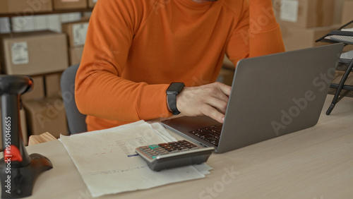 Man holds head and writes on invoice with pen beside laptop and calculator amid stacked cardboard boxes and barcode scanner in building; stress finances.