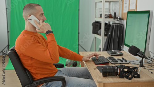 Wallpaper Mural Man photographer holding smartphone to ear while typing with right hand at desk with camera and microphone in studio; focused productivity. Torontodigital.ca